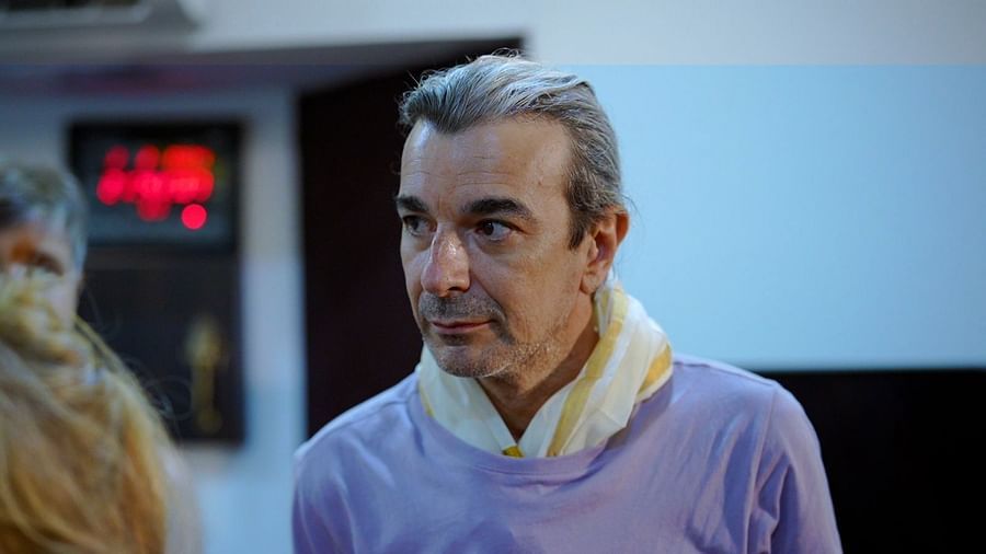 A man with gray hair and a light purple shirt stands in a room, looking slightly to the side, with parts of two other people visible in the foreground.