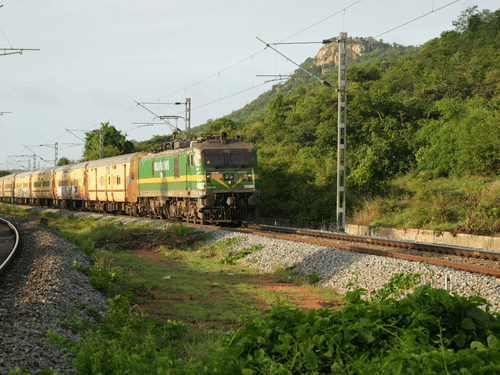 A long passenger train with green and yellow coaches travels along a curved track through a lush green landscape with a large hill in the background.