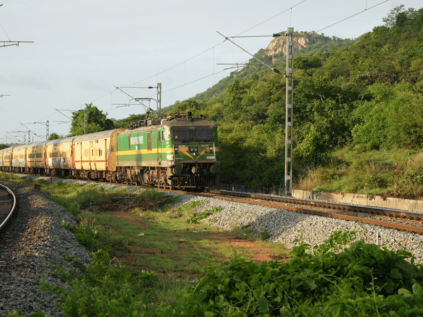 A long passenger train with green and yellow coaches travels along a curved track through a lush green landscape with a large hill in the background.