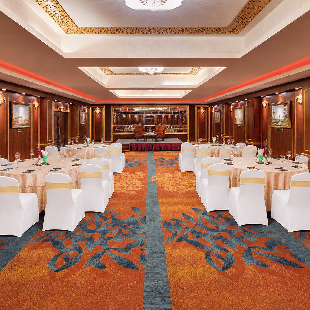 Tulip banquet hall with round tables, white chair covers, patterned carpet, wood-panelled walls with framed paintings and a stage at MAYFAIR Garden, Rourkela.