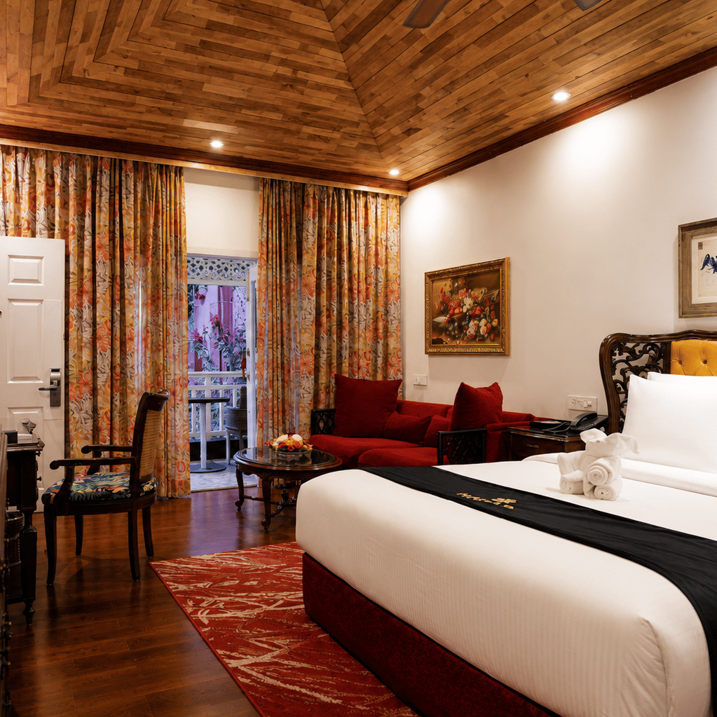 Deluxe Cottage at MAYFAIR Himalayan Spa Resort Kalimpong featuring a bed in the foreground. A sofa sits near a door that opens to the outside. Framed pictures hang on the walls. The ceiling is clad in wood.