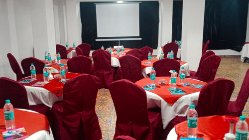 Hall with several round tables set with chairs, displaying a water bottle, book, pen, and a flower vase