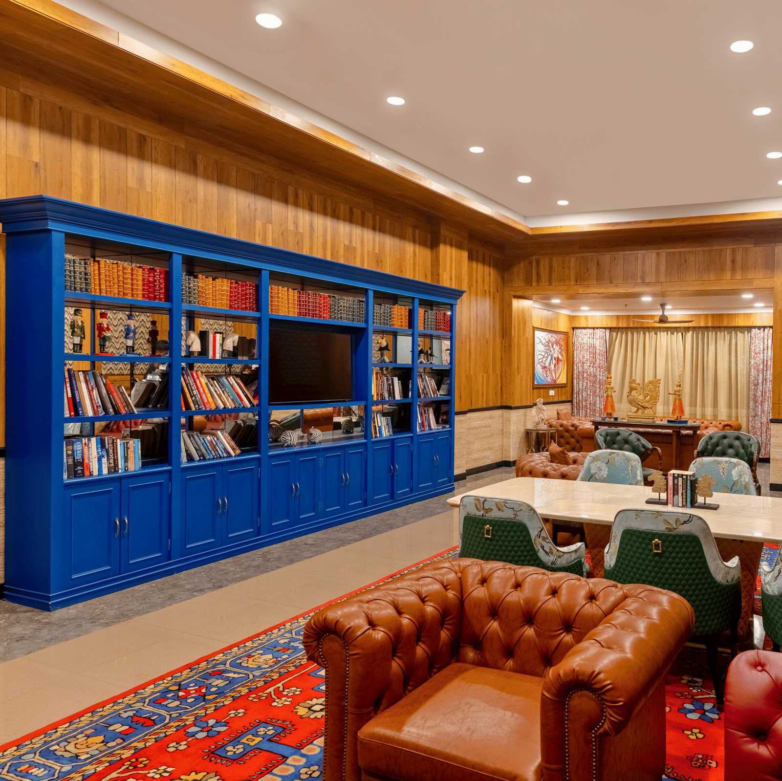 Library lounge with a blue bookcase, leather sofas, upholstered chairs, a marble table and a patterned rug at MAYFAIR Oasis Resort & Convention, Jharsuguda.