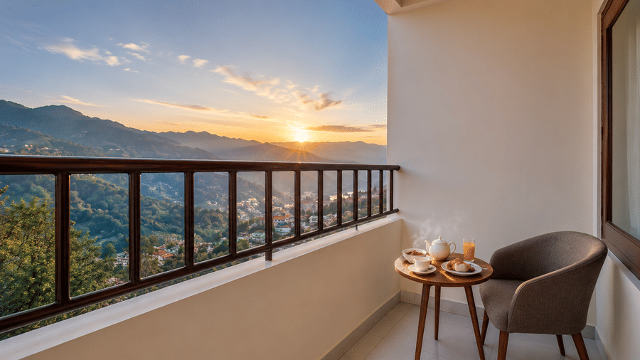 Balcony at Valmere Himalayan Hotel & Spa, Gangtok set with a teapot, breakfast dishes, and an armchair, overlooking a sunrise view of the town and mountains.