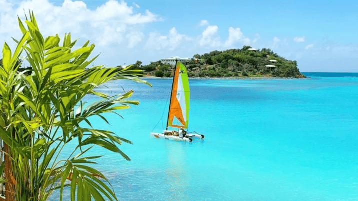 a vibrant tropical scene at Tamarind Hills Resort and Villas in Antigua, featuring turquoise waters, lush green foliage, and a colorful sailboat. 