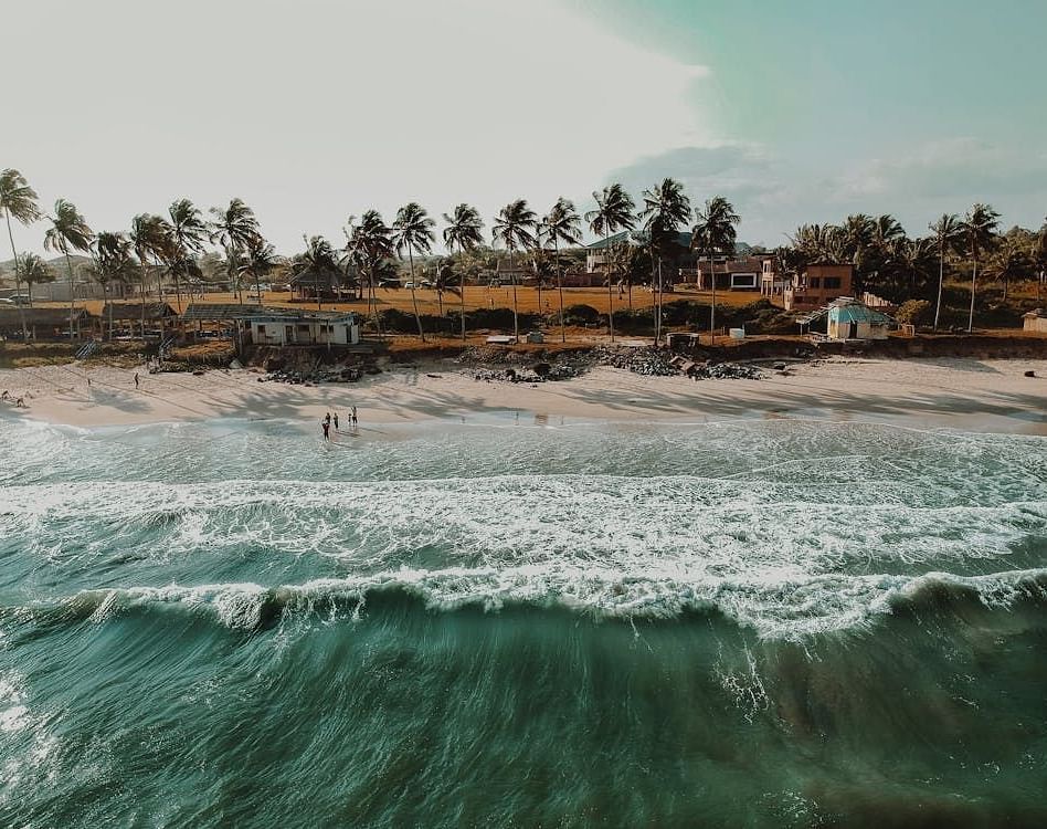 Waves crashing onto a palm-lined beach with fishing boats and structures in the background.