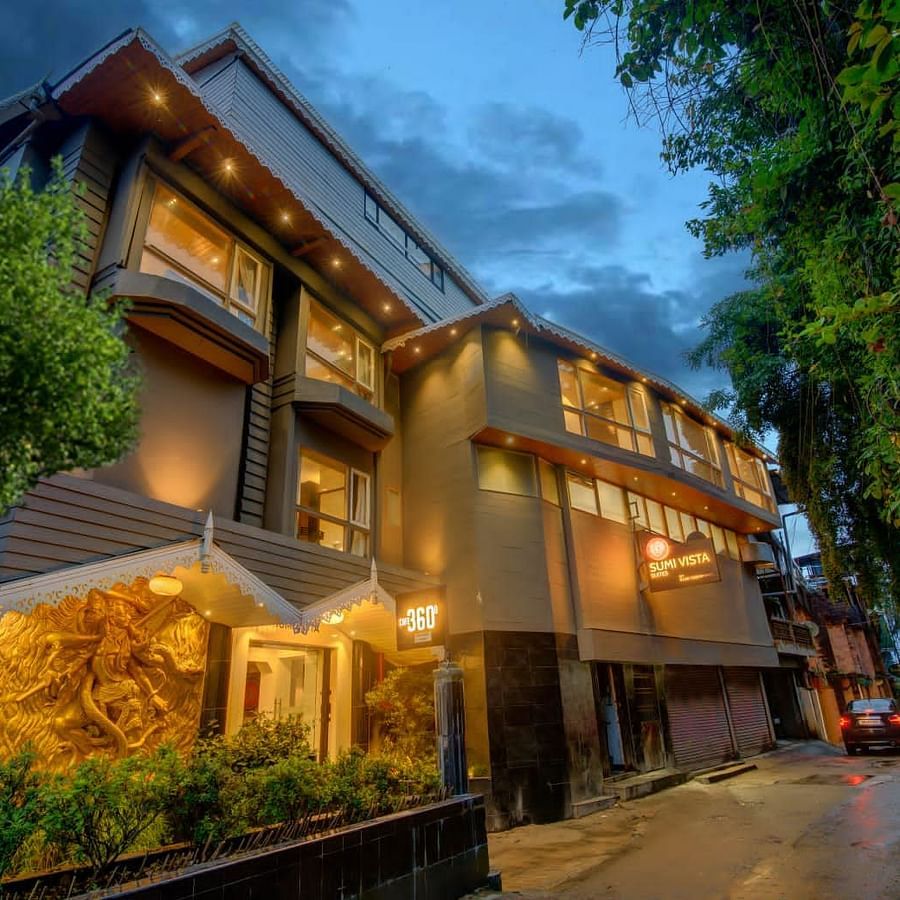 An image of a well lit facade surrounded by trees and there is also a driveway near the entrance - Sumi Yashshree Hotels & Resorts