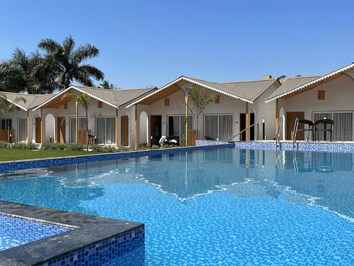 A view of the swimming pool with various accommodations next to the manicured lawn and trees in the distance at VITS Daman Devka Beach - A resort in Daman near the beach.