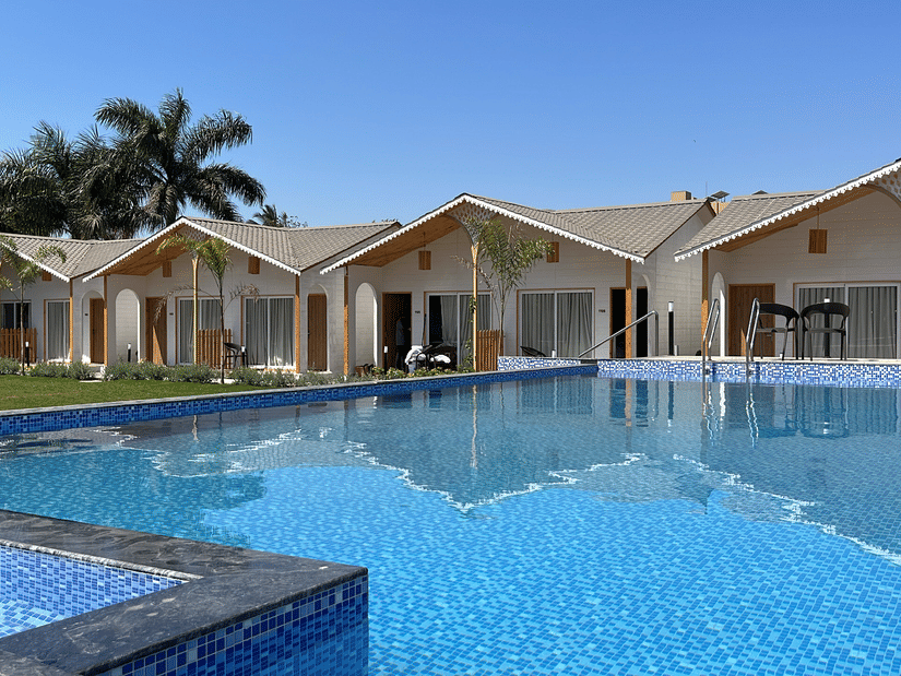 A view of the swimming pool with various accommodations next to the manicured lawn and trees in the distance at VITS Daman Devka Beach - A resort in Daman near the beach.