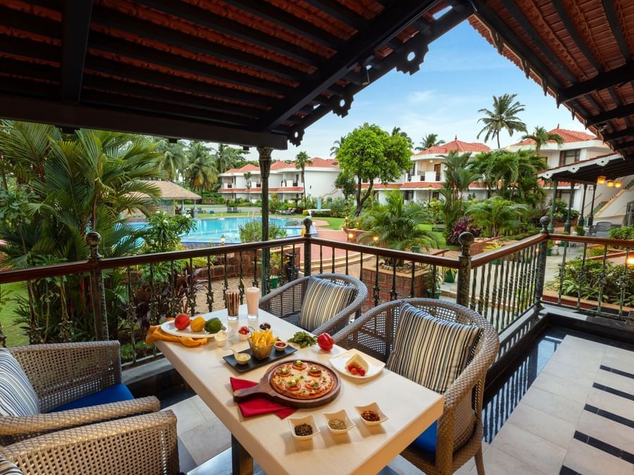 Outdoor dining area with food and drinks on a terrace overlooking palm trees at Heritage Village Resorts & Spa, Goa.