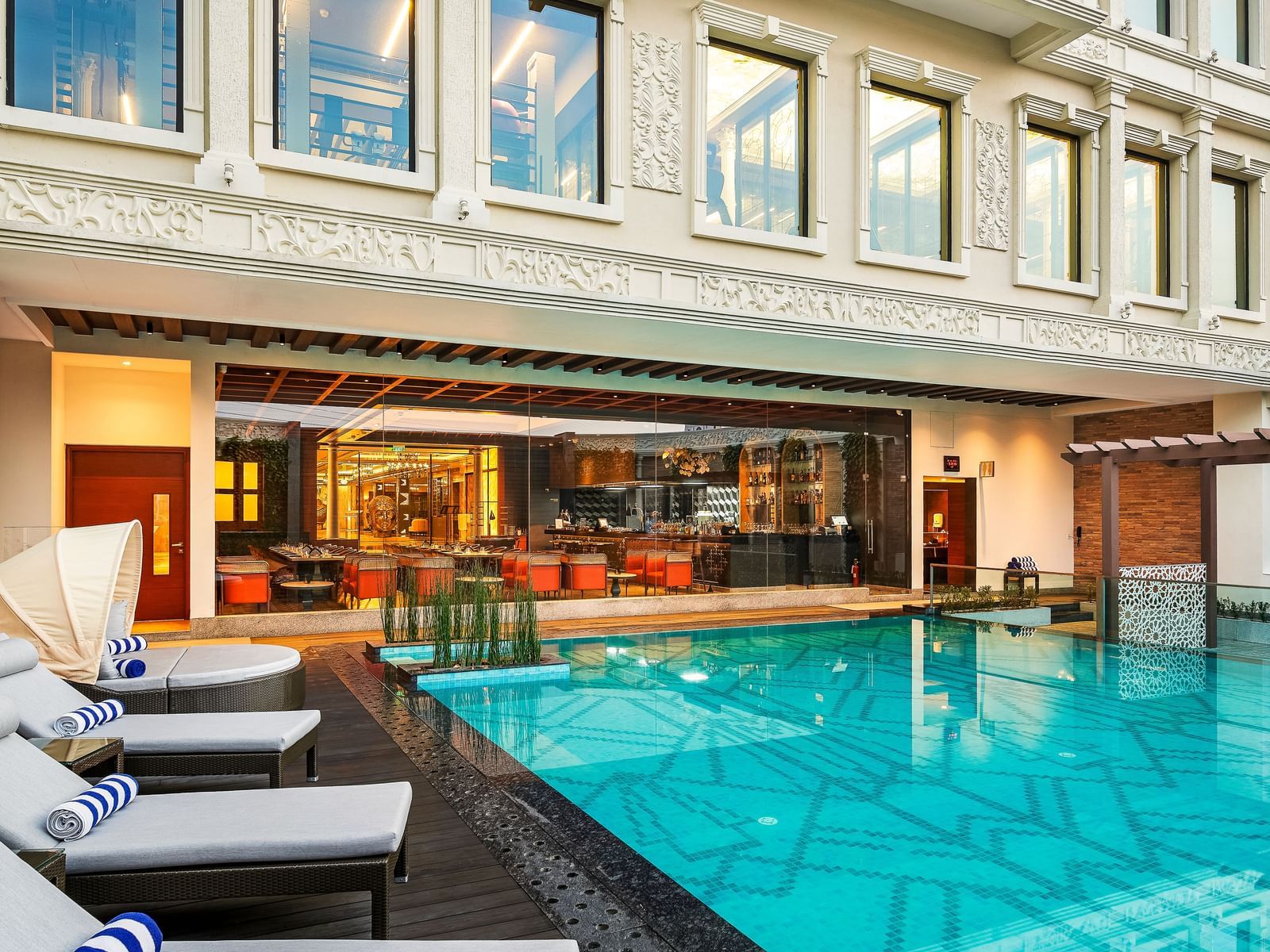 Outdoor setting of Radisson Blur Hotel & Resorts, Chennai, showcasing its swimming pool, lounge chairs, and lobby area as seen through the big glass windows.