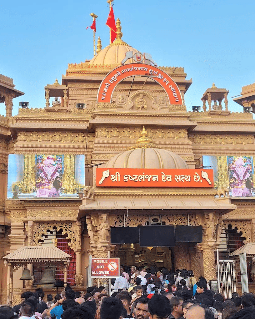 Crowds gathered at the ornate entrance of a Hindu temple with intricate carvings, domes, and digital displays of deities.