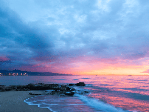 Purple and pink sunset over a rocky beach with gentle waves and a distant city skyline under a cloudy blue sky.
