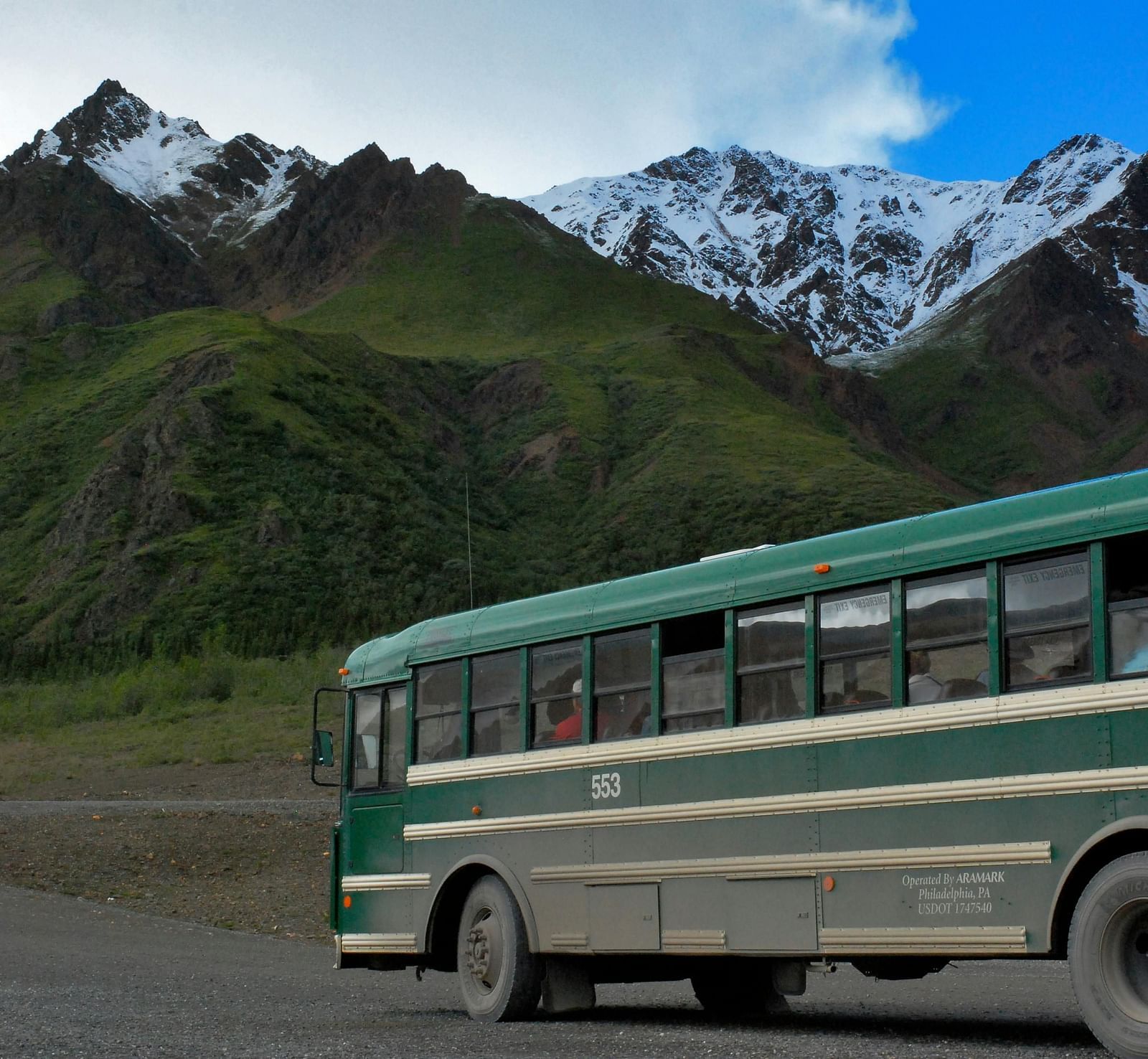 Green bus travelling through a road with green mountains in the background and snow on peak of the mountains.