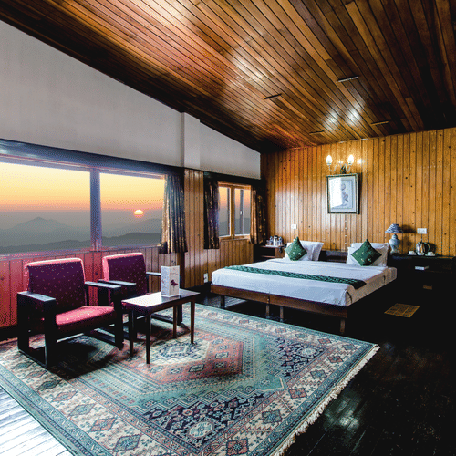 An image of a cosy room with warm wooden interiors, featuring a neatly arranged bed, sofa chairs, and a table, complemented by large glass windows offering a beautiful view of the mountains and the sun - Central Gleneagles Heritage Resort, The Mall Road, D