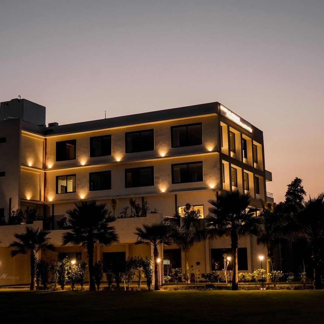 The hotel building of Arawali Ananta Elite Jaipur is illuminated at night with palm trees in front.