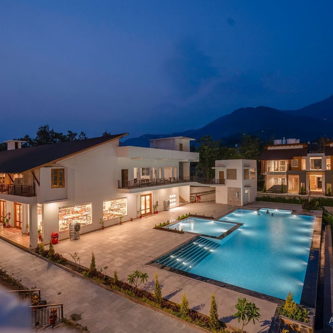 A resort with multiple buildings, a swimming pool, and a large lawn, illuminated at night against a backdrop of mountains.