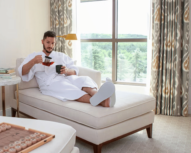  A man in a robe sips from a cup while relaxing on a chaise lounge in a bright room with a large window at YO1 Longevity & Health Resorts
