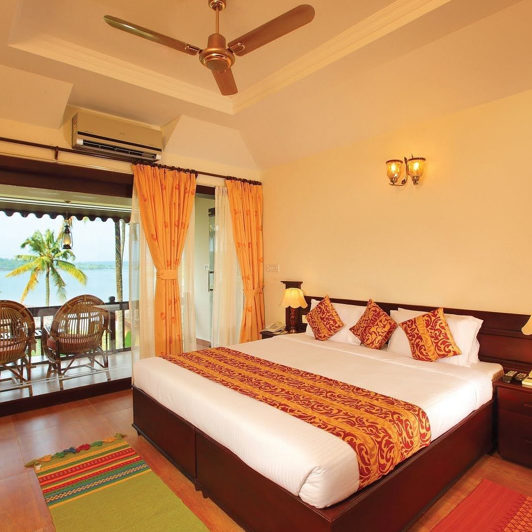 An image of Premium lake View Villa with Private Terrace featuring a double size bed, a bedside table with lamp, and a balcony attached which has two chairs placed - Fragrant Nature Kollam