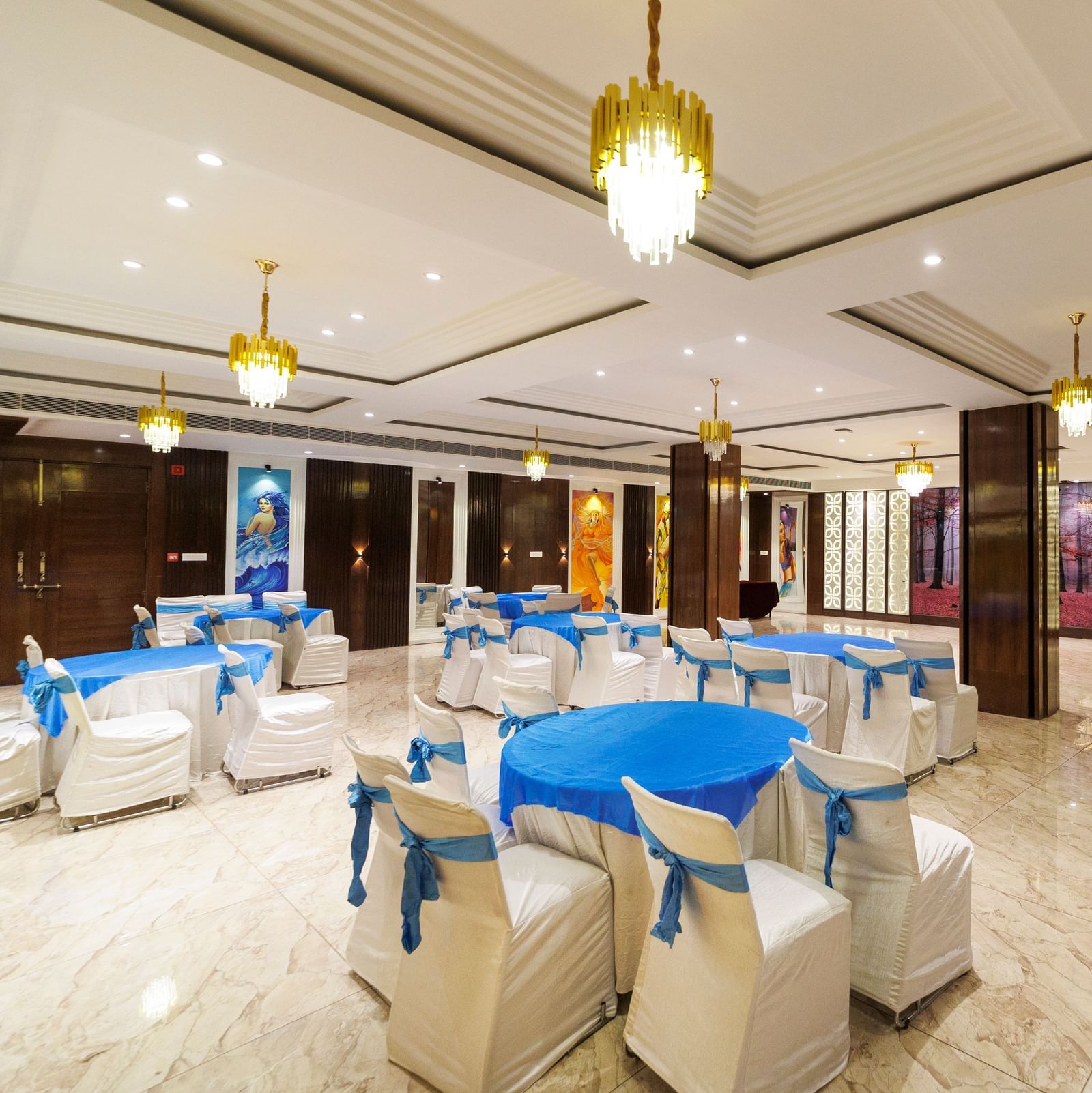 Banquet hall at Inde Reva Resorts Rishhikesh set up with round tables, white chairs, blue covers, and ceiling chandeliers.