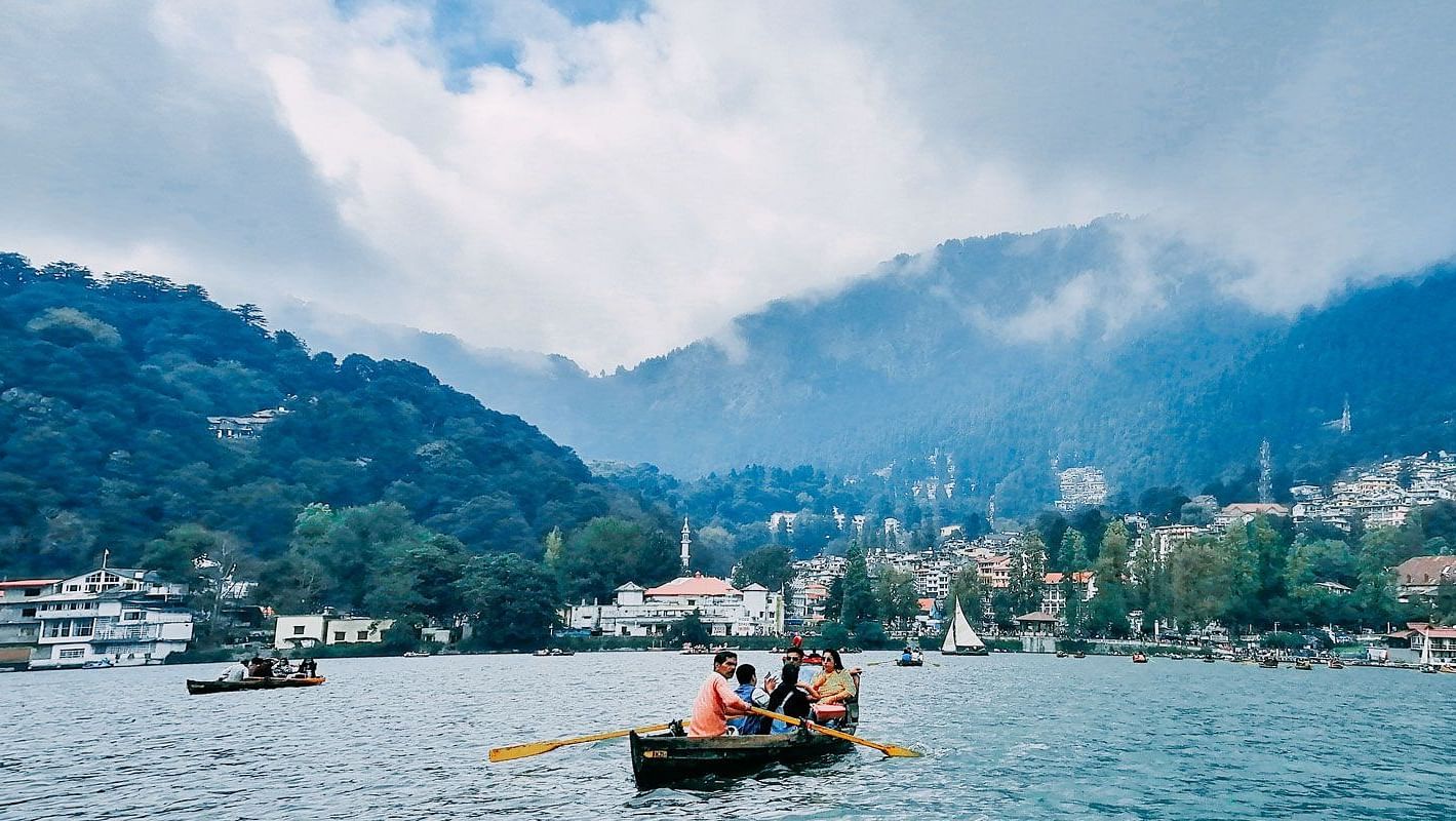 Tourists boating on the tranquil blue waters of Nainital Lake, with forested hills and scattered buildings in the background under a misty sky.