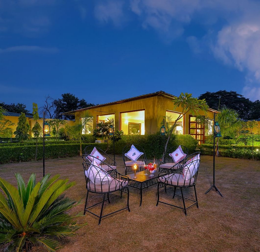 An outdoor dining setup at night featuring a table with four chairs, elegantly set with napkins and drinks, surrounded by greenery and illuminated by soft garden lighting, with a warmly lit building in the background under a deep blue sky - Singhvilas, Ran