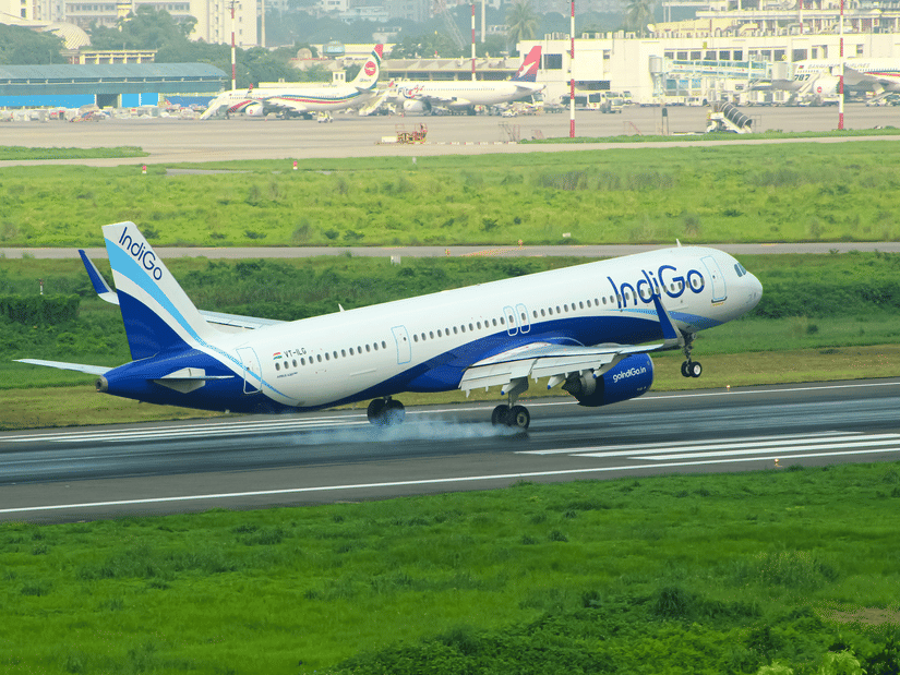 An IndiGo on runway with green grass surroundings, airport buildings in distance, and clear daylight sky