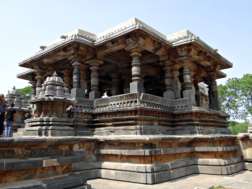 A side view of the Hoysaleswara Temple made of soapstone with pillars in view, people walking near the temple and trees in the background.