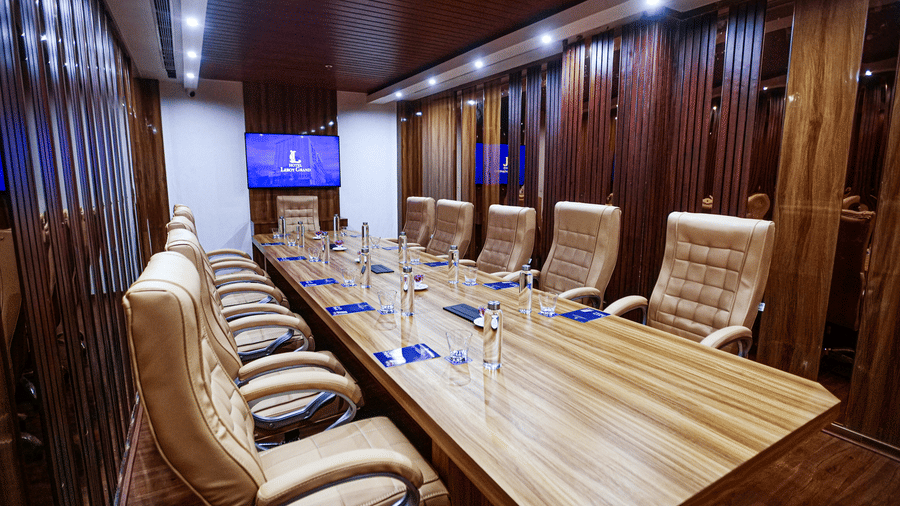 Corner view of the boardroom featuring a long wooden table surrounded by tufted leather cushioned chairs with a wall-mounted television at the far end of the room at Leroy Grand, Yeshwanthpur.