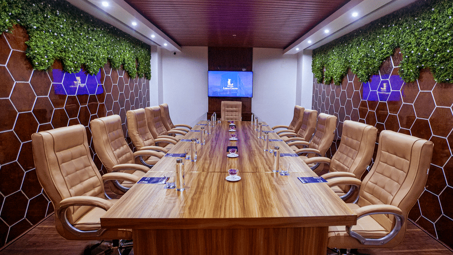Front view of the long wooden conference table placed at the boardroom at Leroy Grand, Yeshwanthpur, with fake green moss used to decorate the walls behind the chairs.