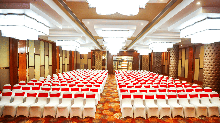 A wide-angle shot of the expansive Mrighashira banquet hall at Leroy Grand, Yeshwanthpur, featuring a carpeted flooring, brightly lit modern might fixtures hanging from the false ceiling, and chairs neatly arranged in lines in theatre setting.