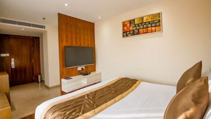 A neatly arranged standard hotel room with a large bed, a wooden headboard, a wall-mounted television, and a colourful painting above the bed at Yashshree The Imperial Inn, Siliguri.