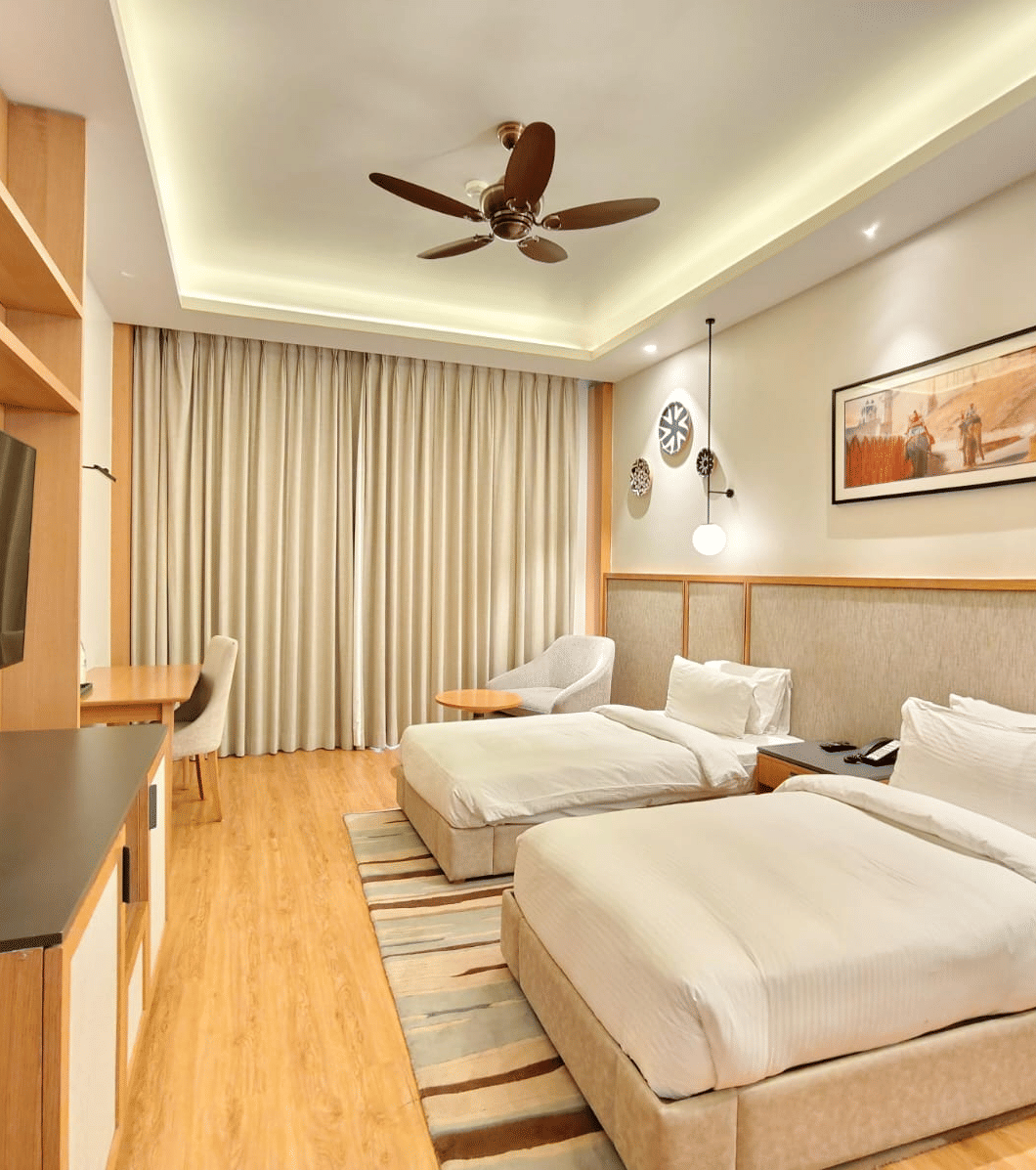 A modern hotel room featuring two neatly made twin beds, warm lighting, a ceiling fan, and a framed picture on the wall.