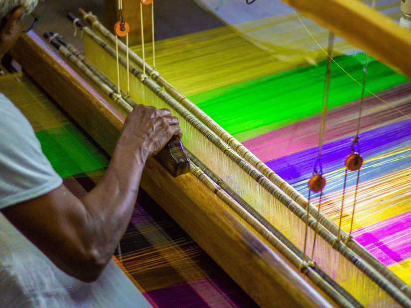 A close up of a person hand weaving a saree with different coloured threads in front of him.