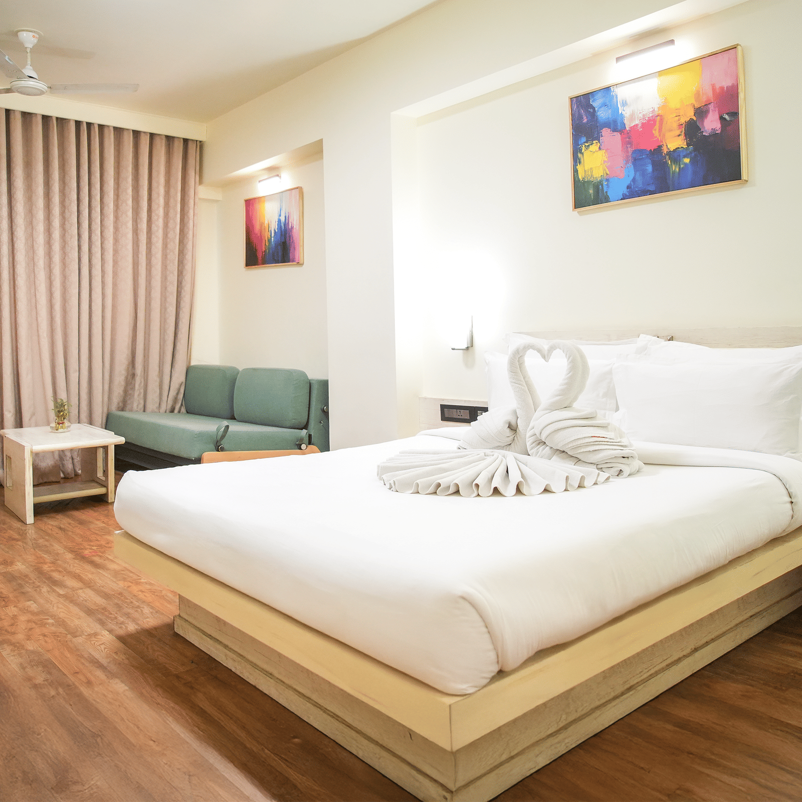 A room with a king size bed, a sofa and a chair, wall arts and light brown curtains at Infiniti Hotel, Indore.