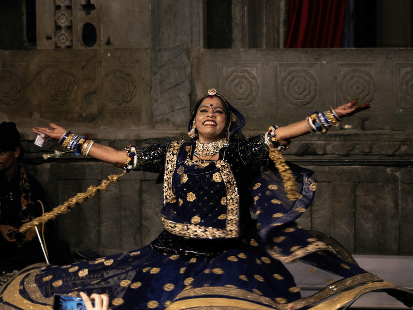 Female folk performer in embroidered costume poses dramatically, illuminated softly against intricately carved stone architecture backdrop
