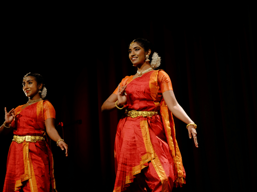 2 dancers performing on stage wearing traditional outfits, with dark background and stage lighting focused on them.
