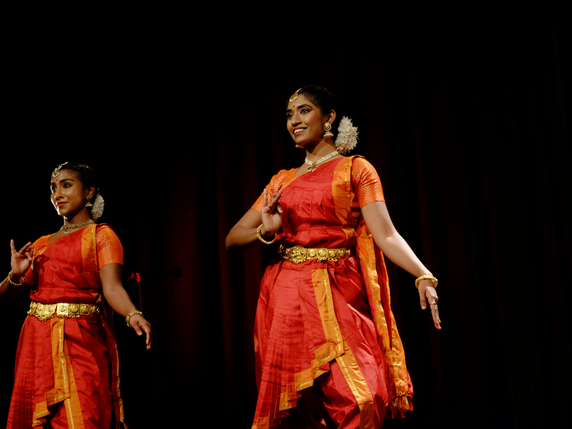 2 dancers performing on stage wearing traditional outfits, with dark background and stage lighting focused on them.