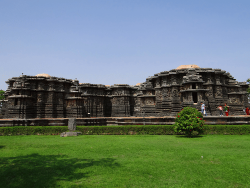 A facade view as seen from a distance of Hoysaleswara Temple with people walking near it and greenery in the foreground.