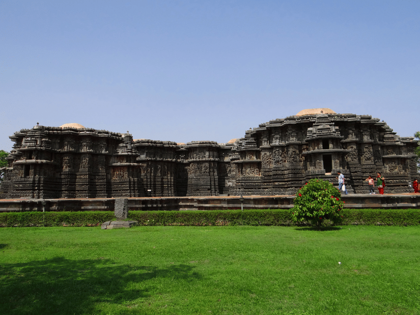 A facade view as seen from a distance of Hoysaleswara Temple with people walking near it and greenery in the foreground.