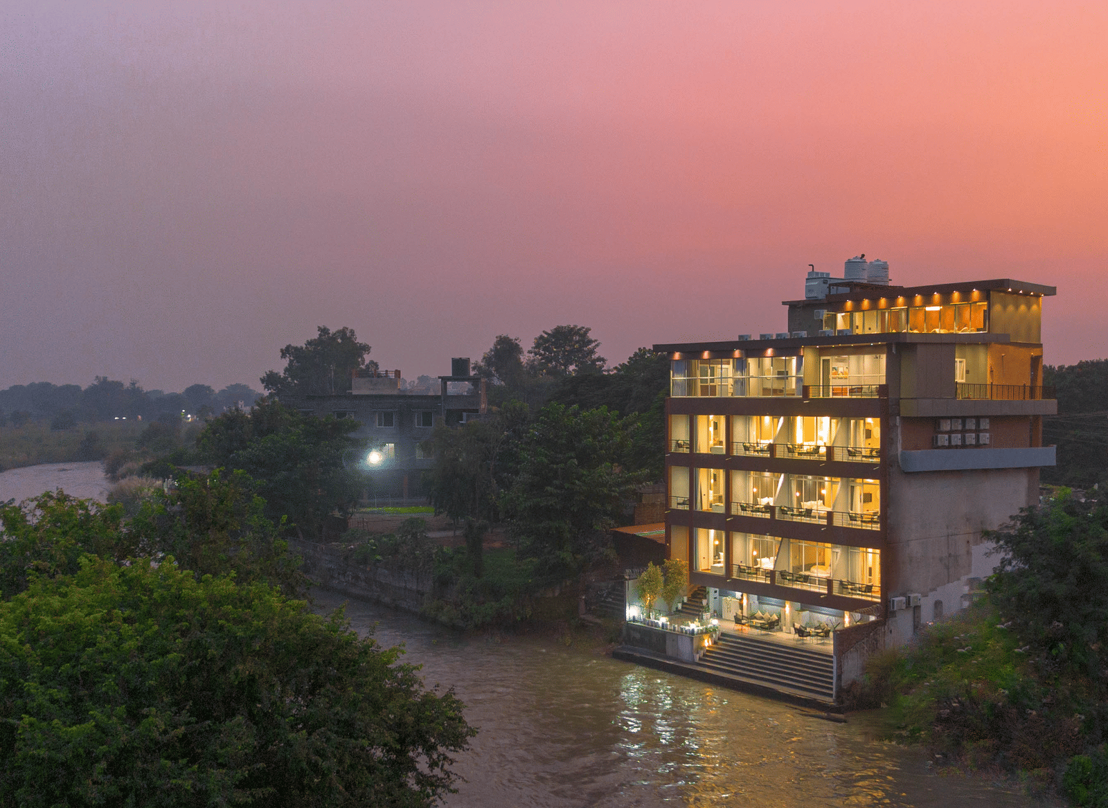 The illuminated Sukoon by the Ganges hotel is captured at sunset, sitting gracefully by the brown-toned river water.