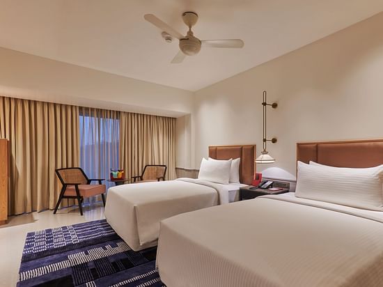 A hotel room with two twin beds, a colorful rug, a dresser, and a view through a window with curtains - The Dukes Retreat