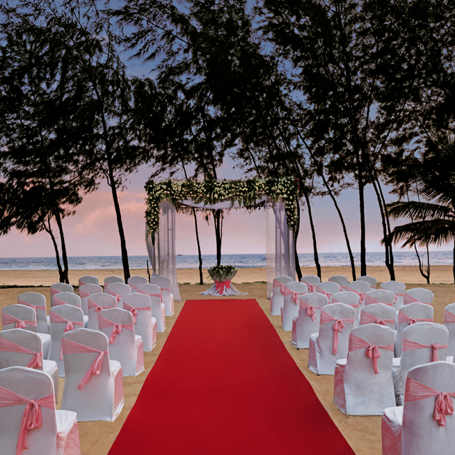 A beach wedding setup at twilight with a red aisle carpet leading to a floral archway, white chairs with pink sashes arranged on the sand, and trees in the background.