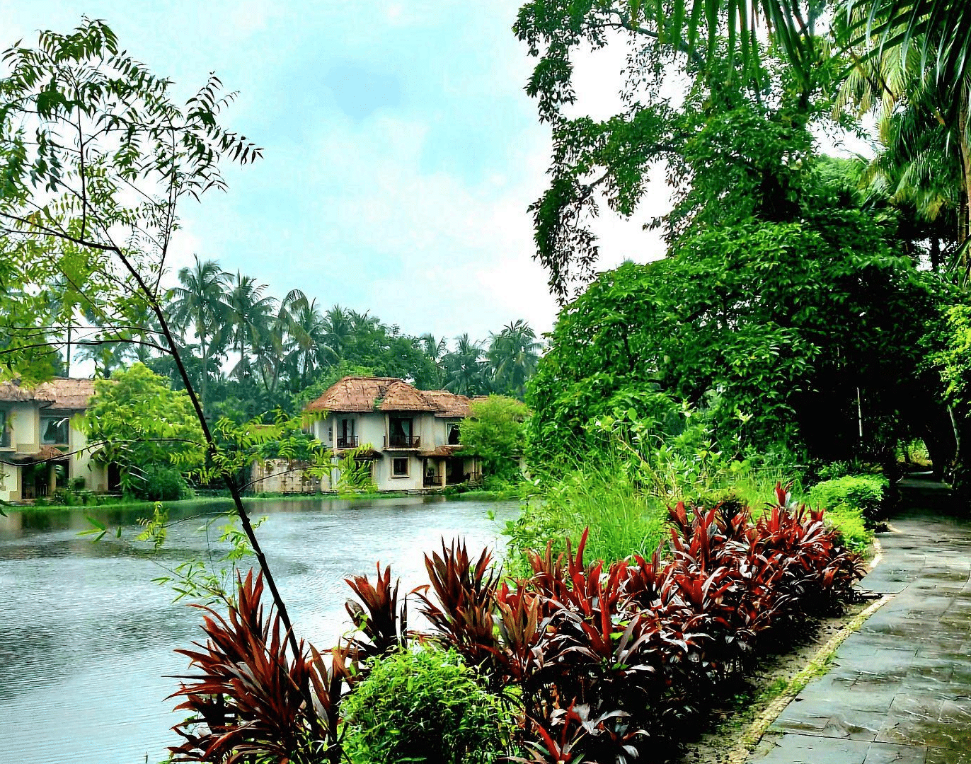 A serene waterside walkway with red and green plants and a fountain, featuring resort buildings in the background at the Vedic Village Spa Resort.