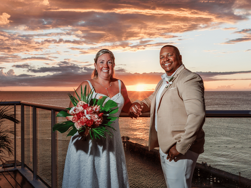 A bride and groom smiling on a balcony overlooking the ocean at sunset, with the bride holding a vibrant bouquet and warm golden light in the background.