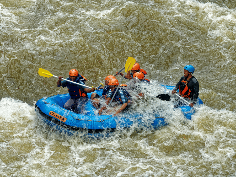 A group of people wearing helmets and life jackets white-water rafting through turbulent river rapids in a blue raft.