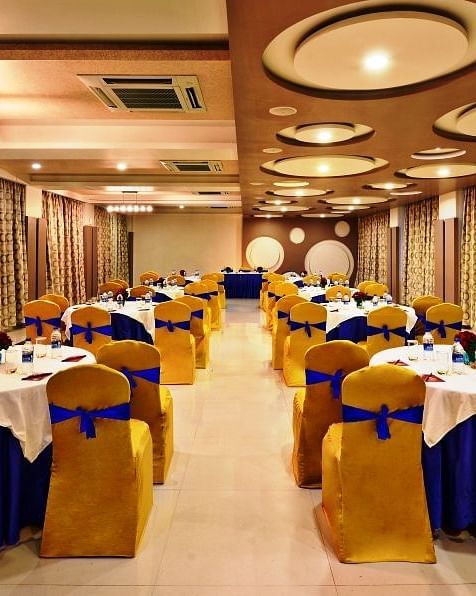 An elegantly decorated banquet hall at Lords Inn Porbandar, with round tables set for dining and chairs with blue and gold accents.