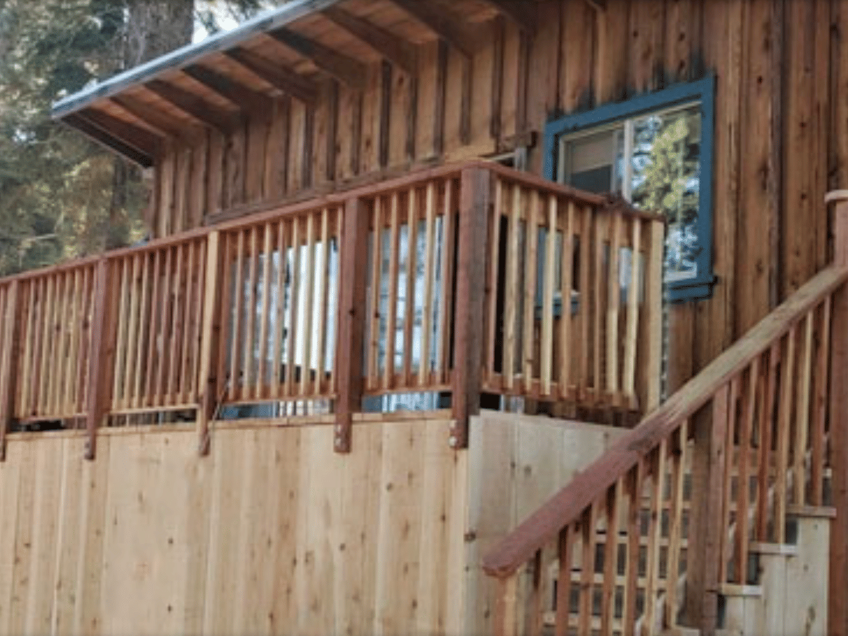A wooden cabin featuring a large deck with a wooden railing and stairs, set amongst trees | Mackinaw