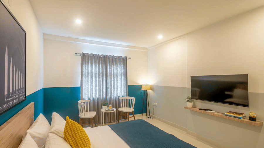A well-lit hotel room at SRS Vivanta Kodihalli featuring a bed with white and teal linens, a wall-mounted TV, and a small seating area by a window with grey curtains.