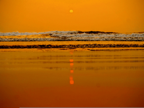A shot from the surface of the shoreline at a beach in Goa showcasing the twilight view of the sun setting in the horizon and the waves crashing on the shoreline.
